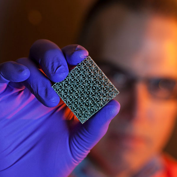 A researcher holds an additively manufactured metal cube up to the camera